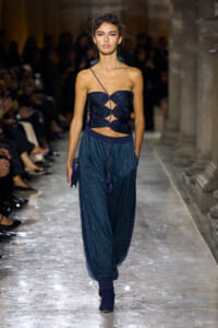 Model walks the runway in a navy glitter crop top with geometric cutouts and matching shimmering jogger-style pants; audience blurred in the background.