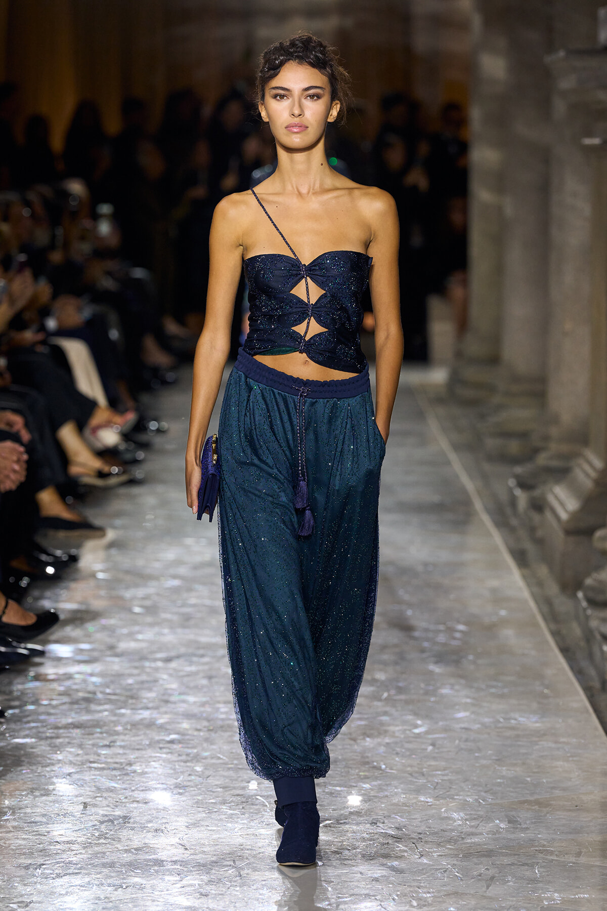 Model walks the runway in a navy glitter crop top with geometric cutouts and matching shimmering jogger-style pants; audience blurred in the background.