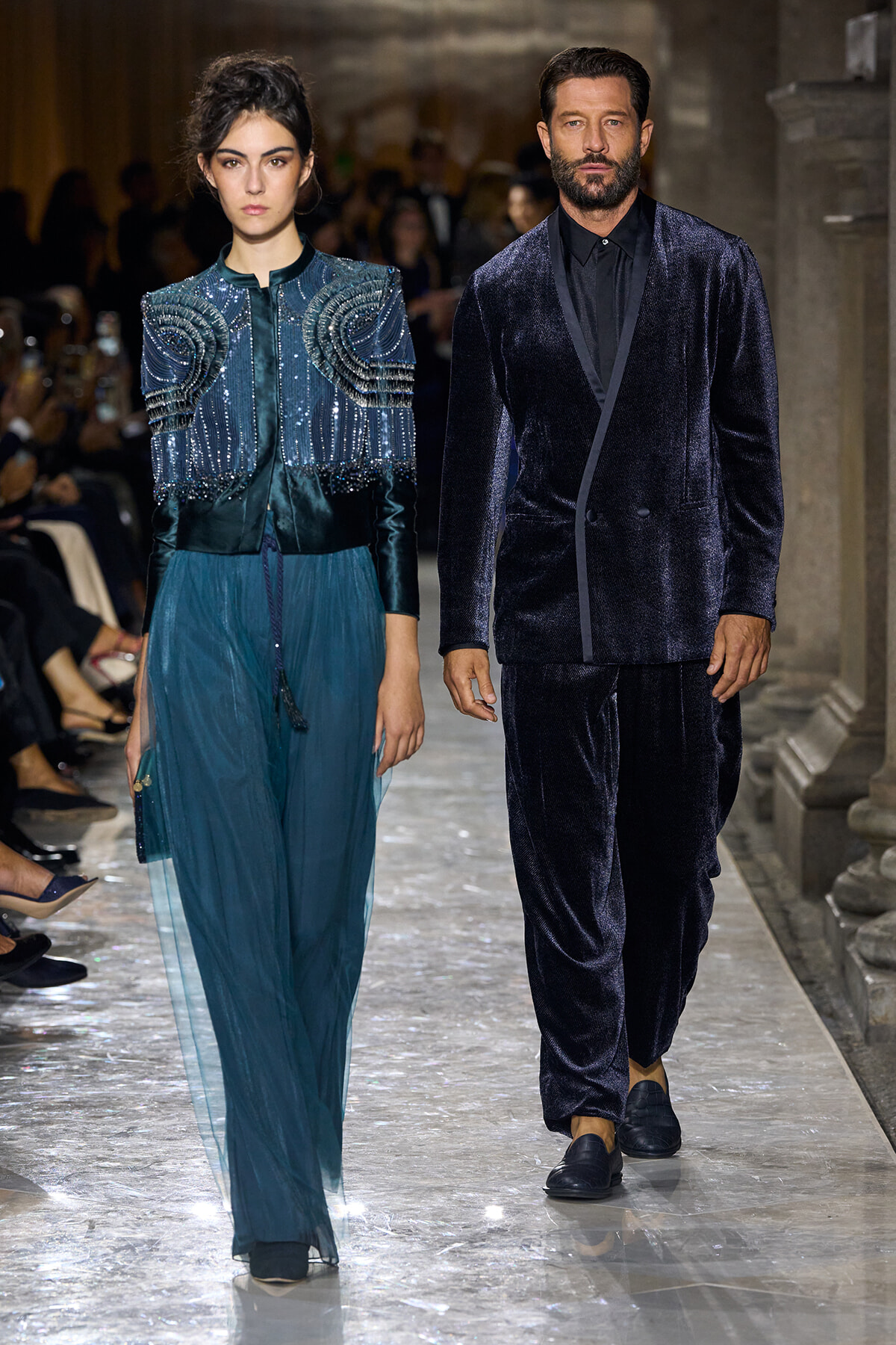 Two models walk a runway in formal teal and navy velvet outfits; the woman wears beaded teal jacket and sheer skirt, the man a dark velvet suit.