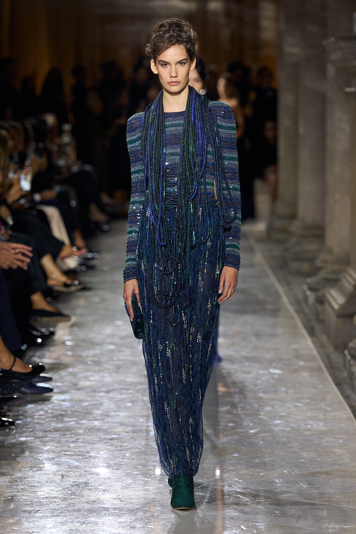 Model walks the runway in a long blue sequined dress with layered blue beaded scarves and a dark clutch.