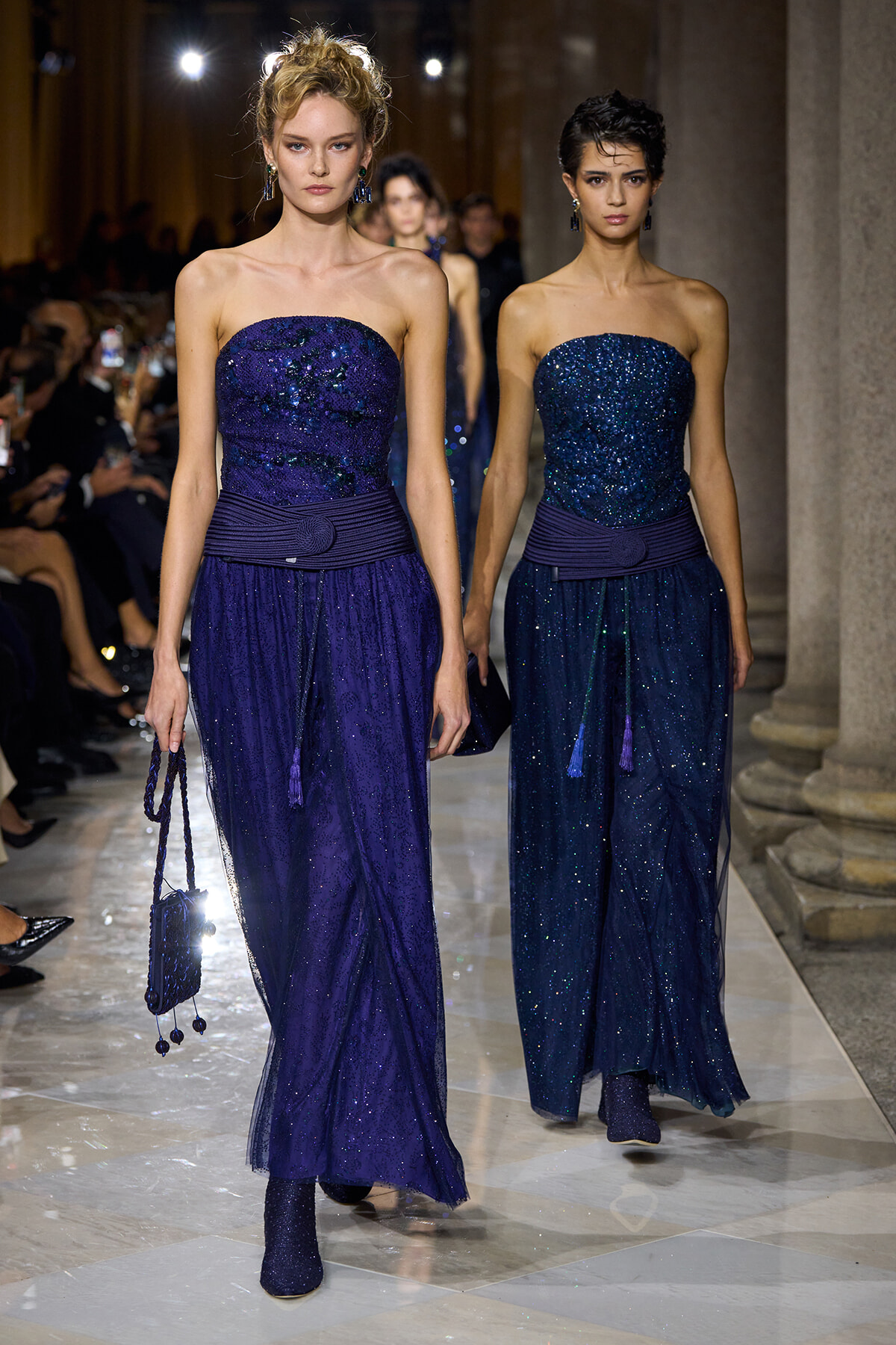 Two models walk a runway wearing strapless royal blue sequin gowns with wide belts and matching accessories.