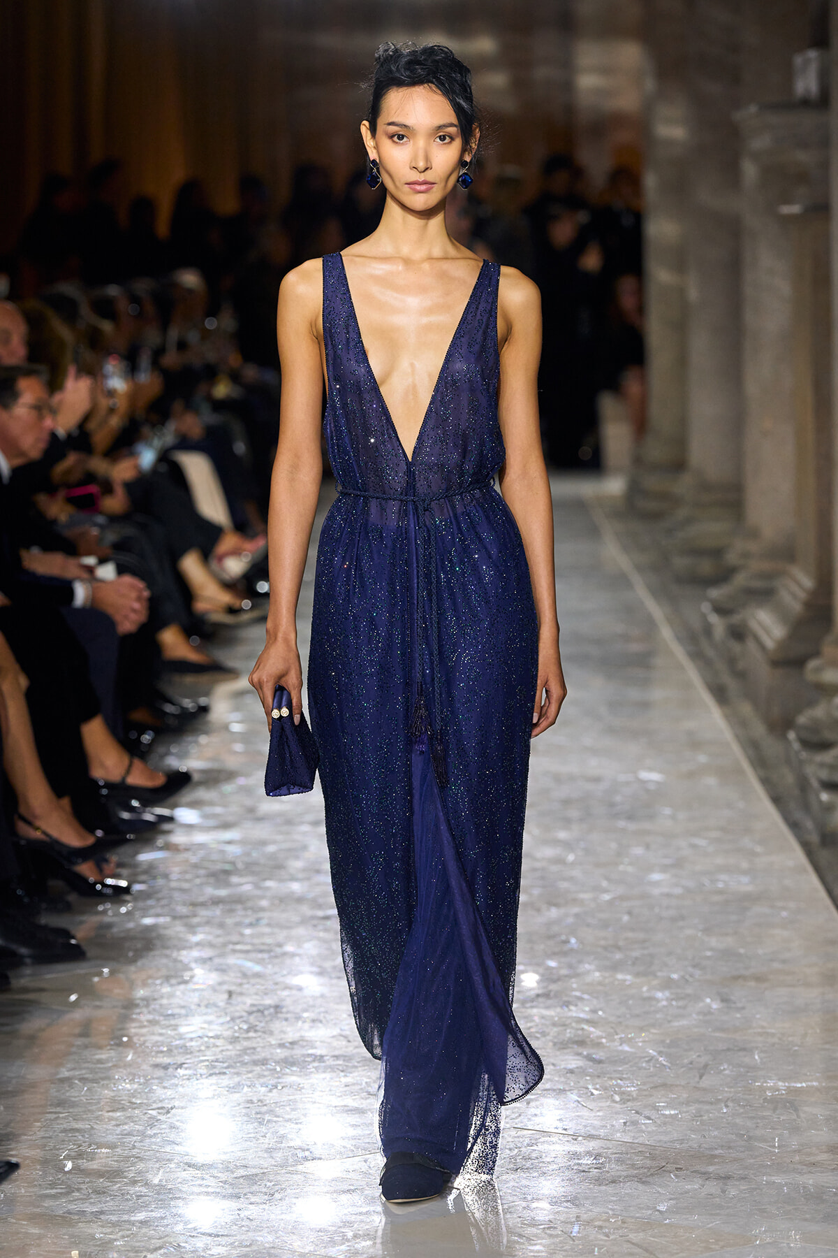 Model walking a sparkly navy blue gown with a deep V neckline on a runway, audience seated along the sides.