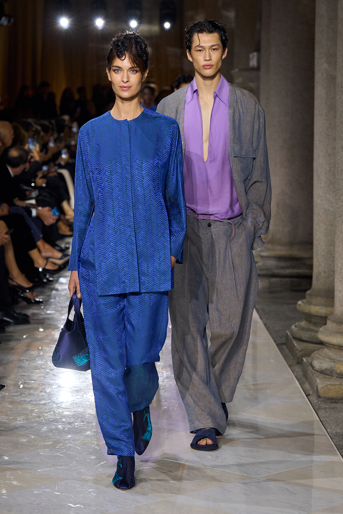 Two models walk a runway; woman in a blue patterned tunic and wide pants carries a blue bag, man in lavender shirt and gray suit jacket follows behind.