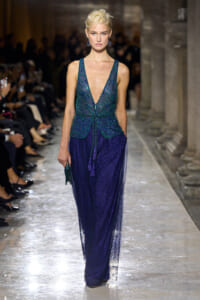 Model walks the runway in a deep V emerald sequin top and sheer navy blue skirt, with statement blue earrings and a small green clutch, in a grand, columned venue.