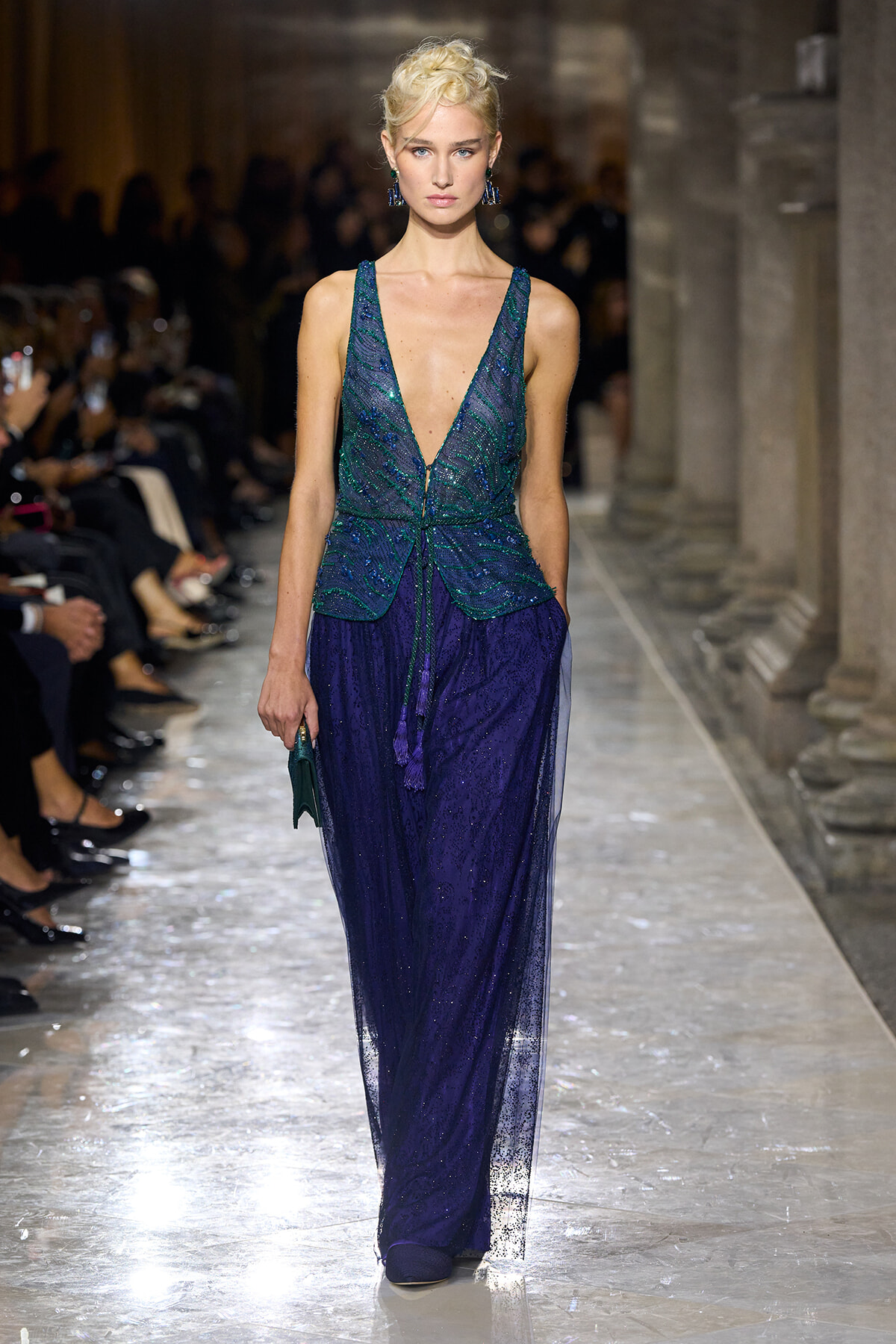 Model walks the runway in a deep V emerald sequin top and sheer navy blue skirt, with statement blue earrings and a small green clutch, in a grand, columned venue.