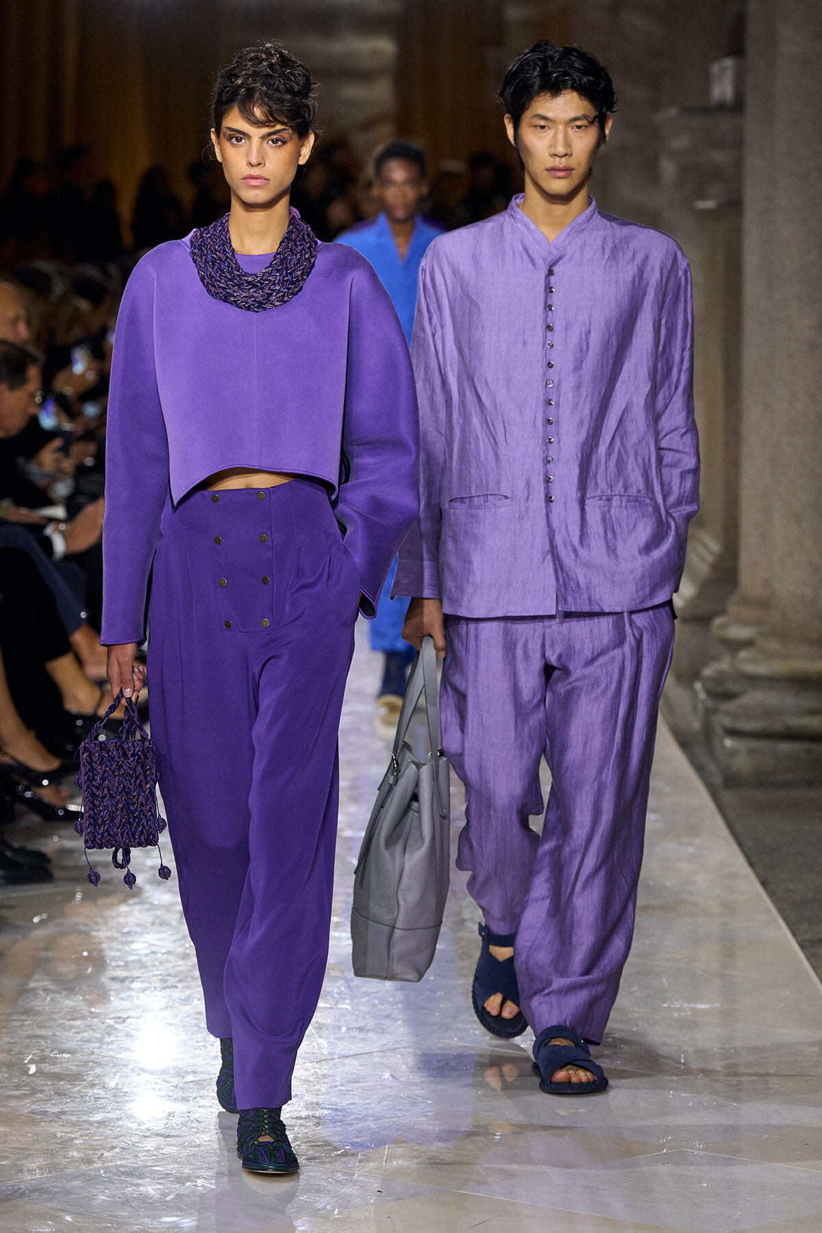 Two models walk a runway in purple outfits: a cropped top and high‑waisted pants with accessories on the left, a lavender linen suit on the right.