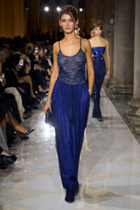 Model walks the runway in a cobalt blue sequined gown, audience members capturing photos on both sides.