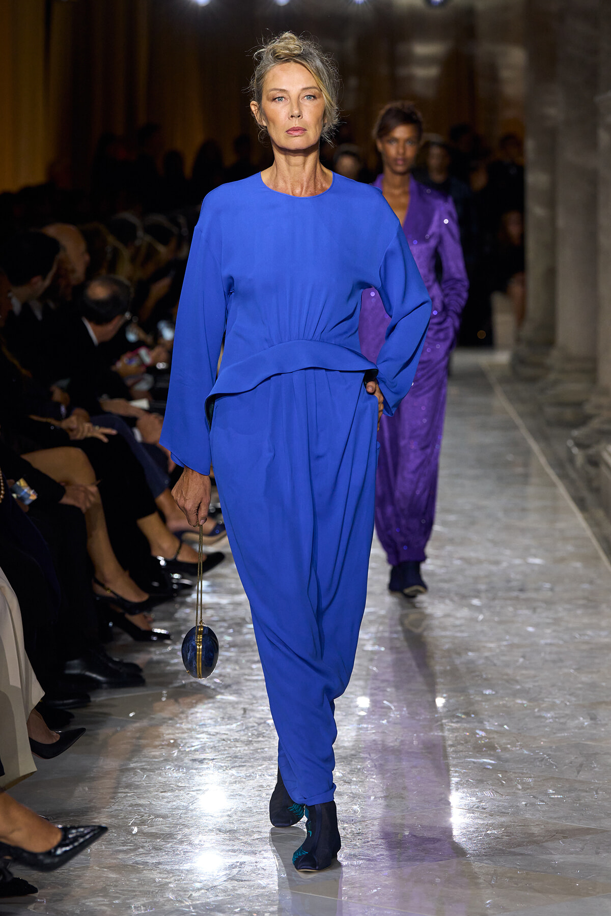 Model in a royal blue belted jumpsuit walks the runway, holding a small round blue handbag on a fashion show.