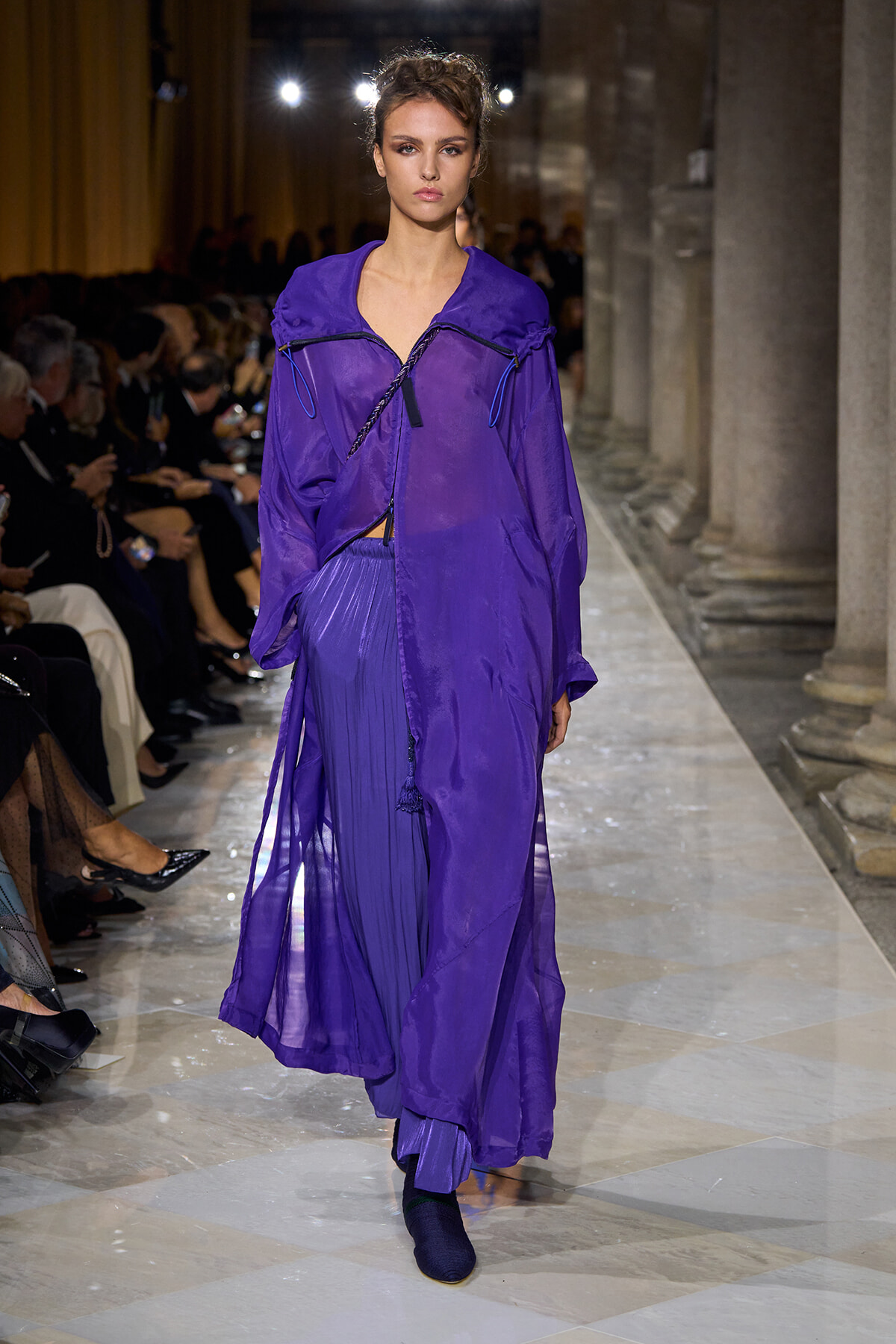 Model walks a runway in a loose, vibrant purple ensemble with a long, flowing cape-like layer and pleated skirt in a fashion show setting.