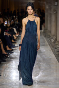 Model walks the runway in a dark blue halter gown with a shimmering gradient, holding a small handbag on a glittering marble catwalk.