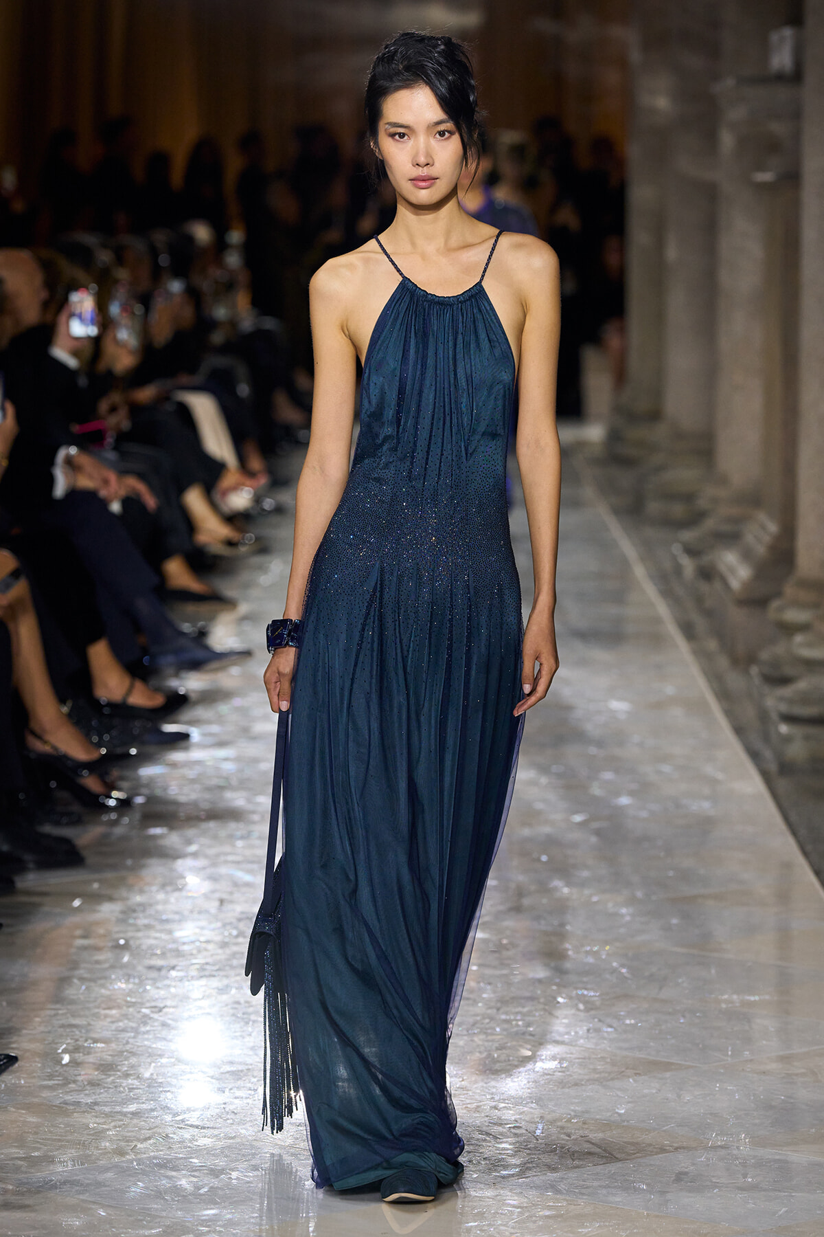 Model walks the runway in a dark blue halter gown with a shimmering gradient, holding a small handbag on a glittering marble catwalk.