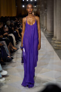 Model walking a fashion runway in a long, cobalt purple halter dress with a draped front, audience on both sides.