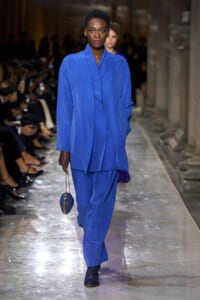 Model walking the runway in a bright cobalt blue layered ensemble, carrying a small blue circular bag with a gold chain and wearing matching blue shoes.