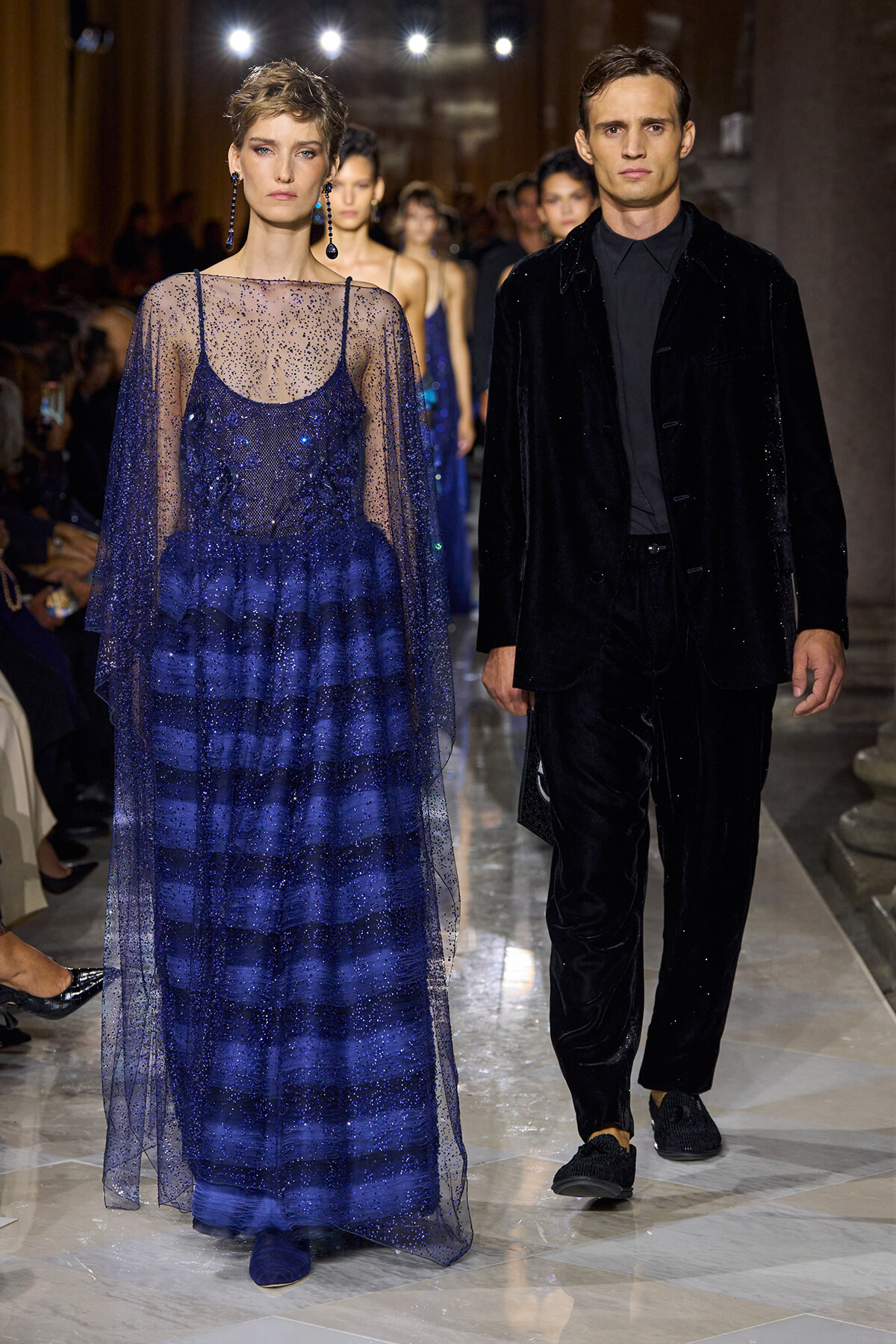 Female model in a sparkling cobalt blue gown with a sheer dotted cape walking a fashion runway beside a male model in a black velvet suit.