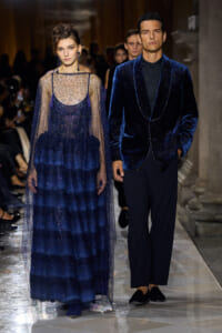 Two models walk a runway in cobalt blue eveningwear: a sheer, beaded navy gown with a layered skirt and a velvet-trimmed cape, and a matching velvet blazer with black pants.
