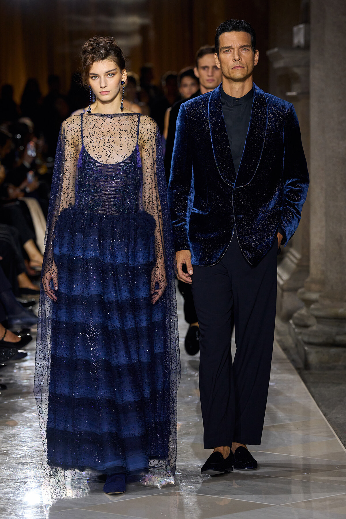 Two models walk a runway in cobalt blue eveningwear: a sheer, beaded navy gown with a layered skirt and a velvet-trimmed cape, and a matching velvet blazer with black pants.