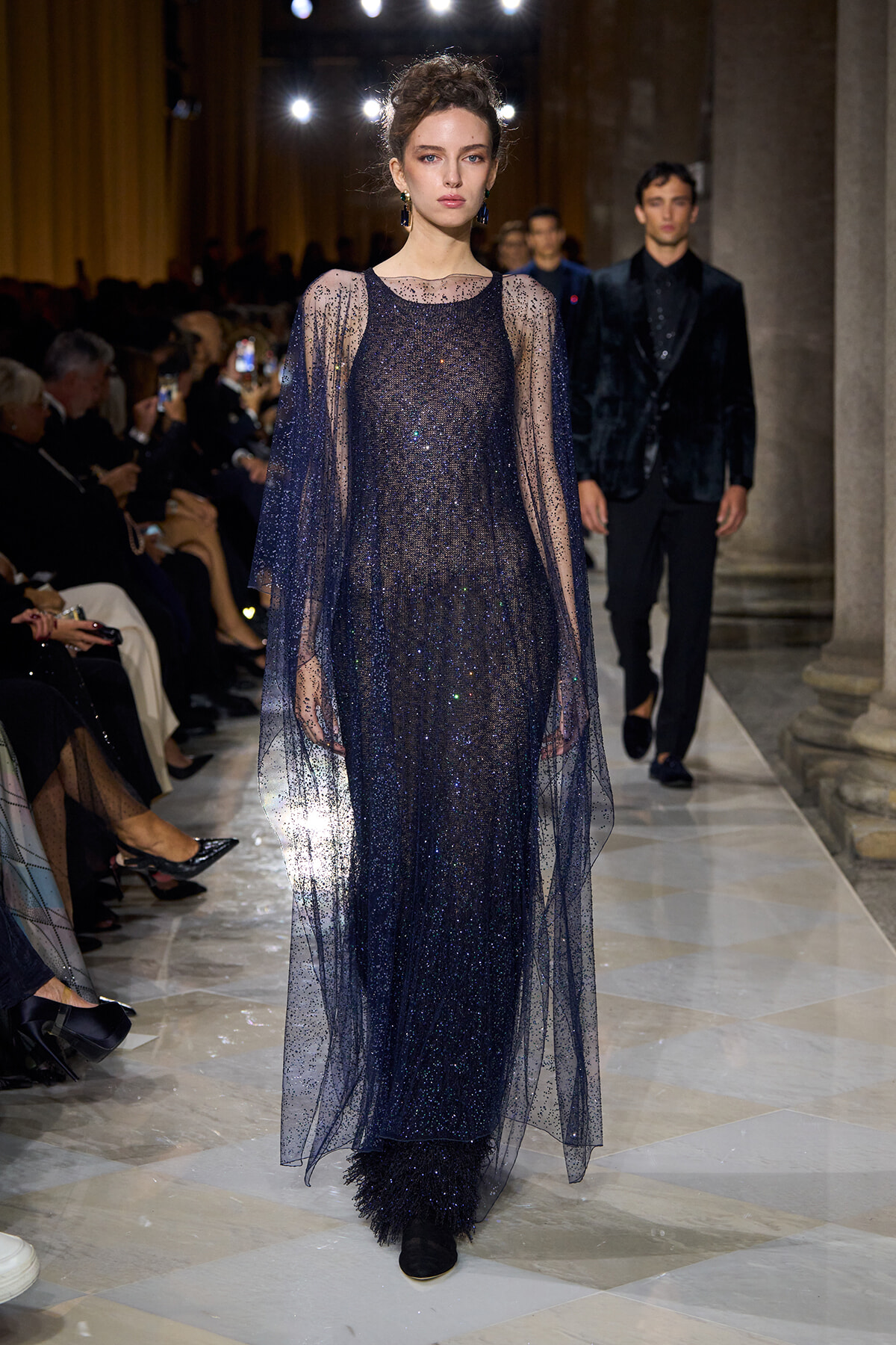 Model walks the runway in a deep navy, glittering gown with a sheer overlay cape and matching accessories.