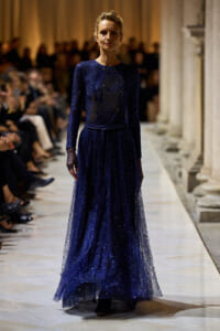 Model walks the runway in a floor-length navy blue glitter gown with long sleeves and a sheer tulle overlay.