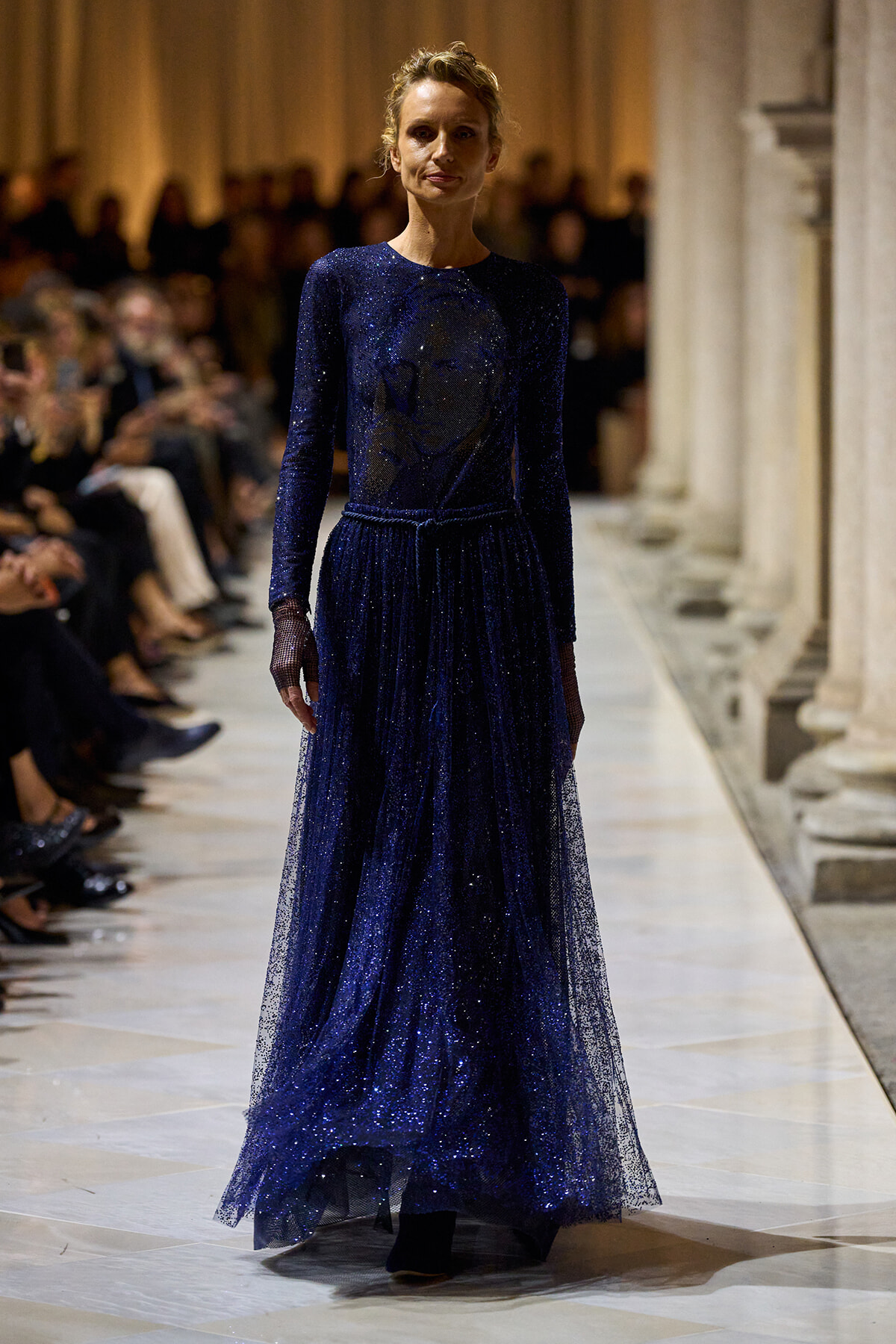 Model walks the runway in a floor-length navy blue glitter gown with long sleeves and a sheer tulle overlay.