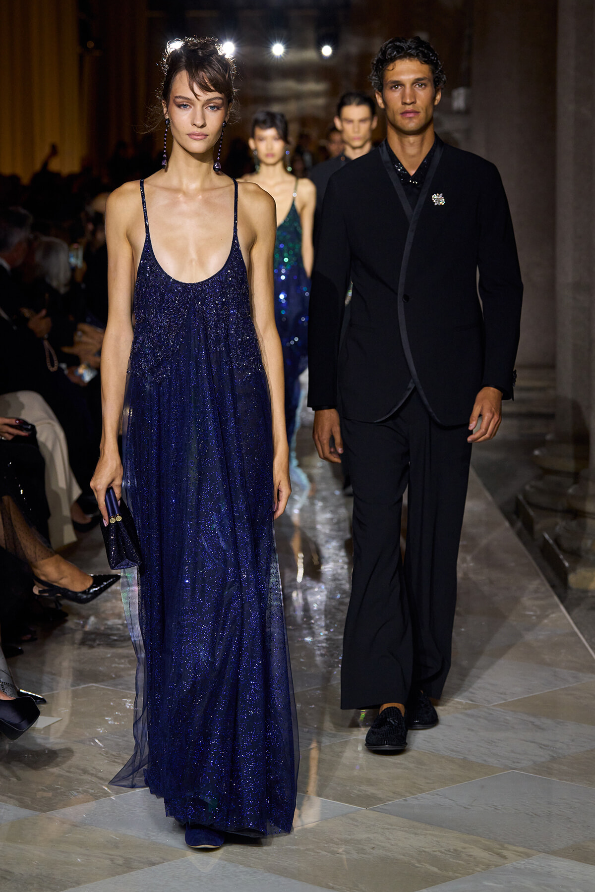 Female model in a deep navy glitter gown with thin spaghetti straps, carrying a matching clutch, walking the runway beside a man in a black suit.