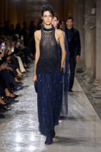 Model walks the runway in a dark blue, beaded evening gown with a sheer bodice and layered skirt, during a fashion show.