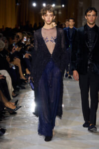 Model walks a fashion runway in a navy sequin gown with a sheer, embroidered bodice and darkness at the edges, audience watching.”,