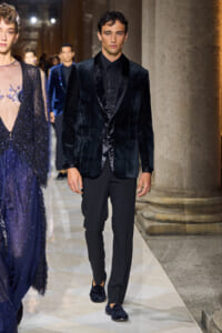 Male model on a fashion runway wearing a black velvet blazer, black shirt, and tailored black trousers; velvet loafers complete the look.