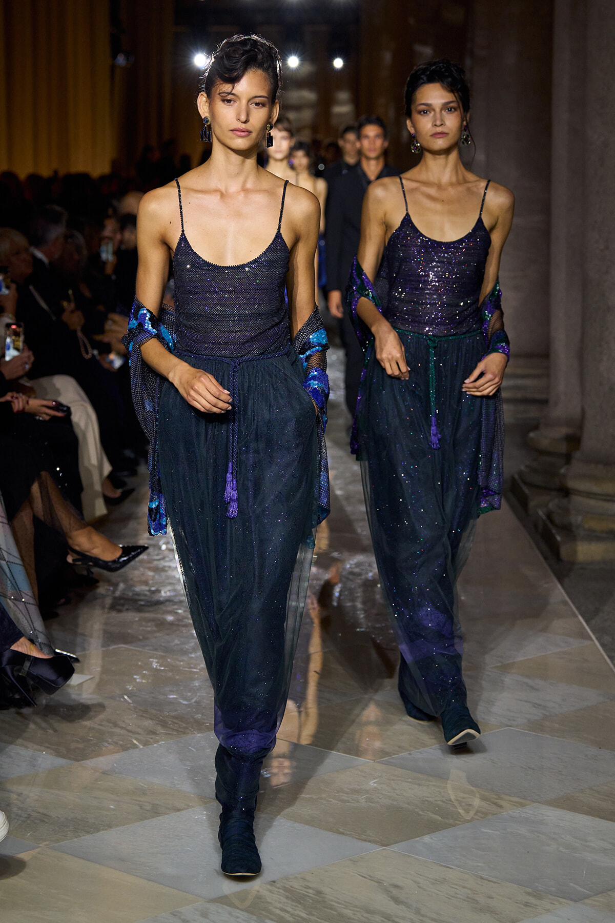 Two female models walk a runway in navy glittery outfits with draped shawls and teal tassels.