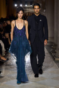 Two models walk a runway: a woman in a blue gradient, sheer evening gown with a purple embellished bodice beside a man in a black suit and vest.