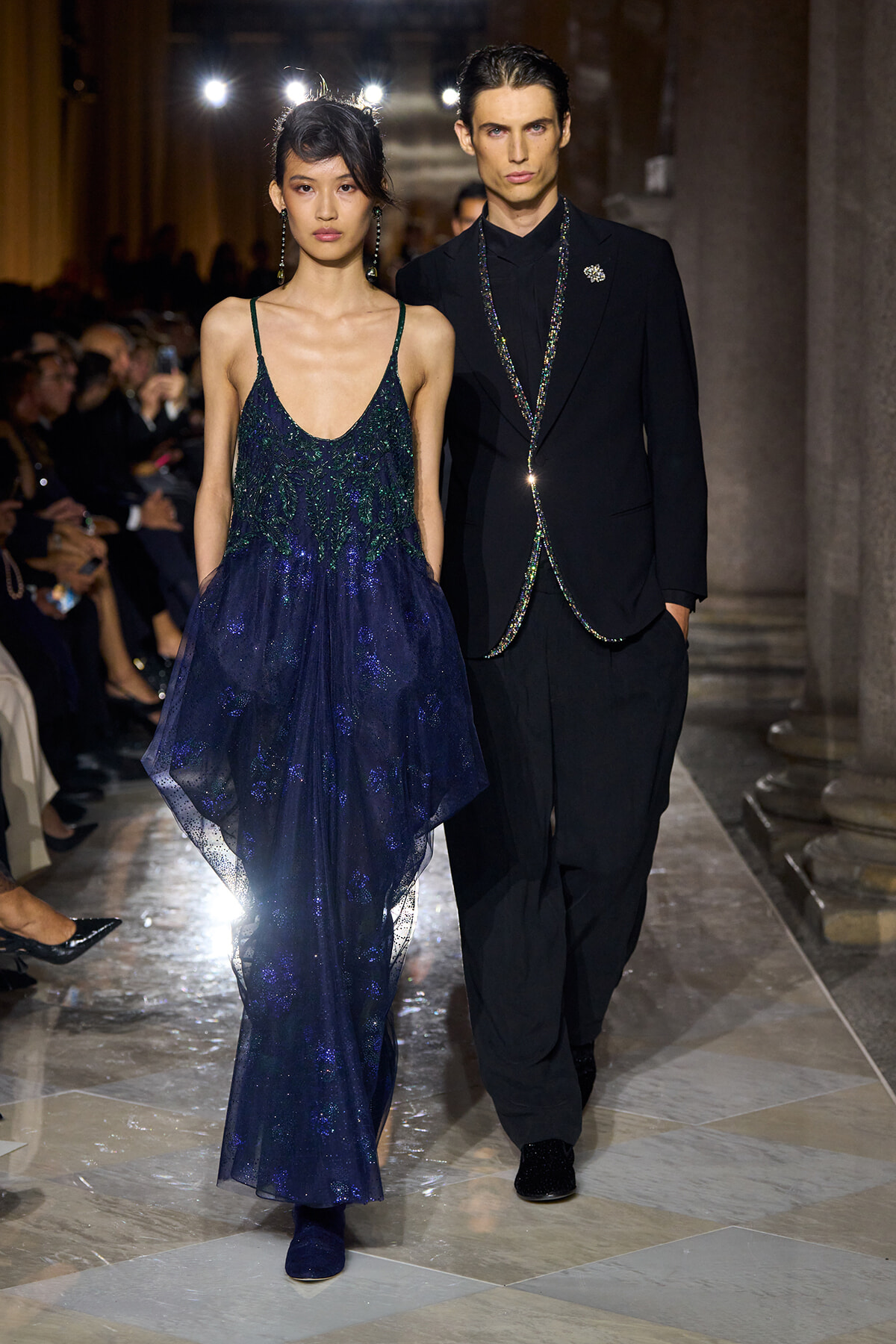 Woman in a navy, sparkly evening gown with thin straps walking the runway beside a man in a black suit as the audience watches.