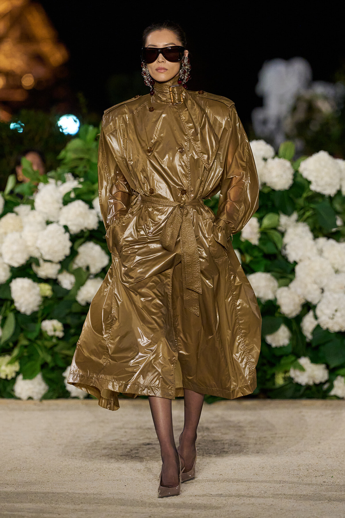 Model walking a runway in a bronze metallic trench coat with belt, oversized sunglasses, and bold earrings, against a backdrop of white hydrangeas at night