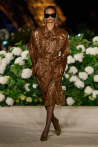 Model on a runway wearing a brown glossy trench coat belted at the waist, oversized sunglasses, and statement earrings, with white hydrangeas in the background.