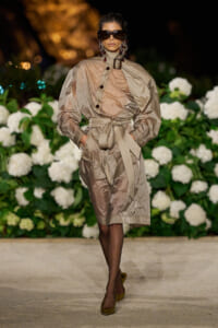 Model walking a runway in a metallic beige trench dress with a wide belt and oversized sleeves, dark sunglasses, and statement earrings.