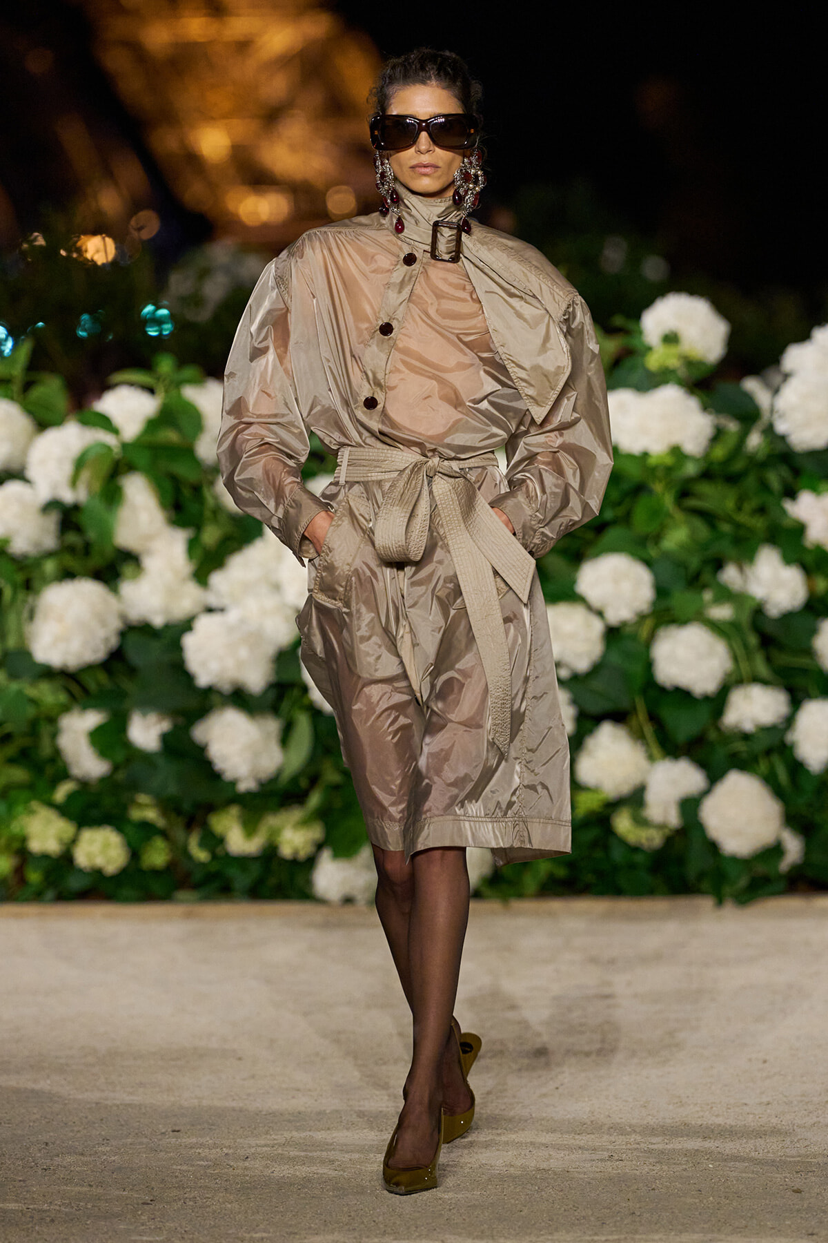 Model walking a runway in a metallic beige trench dress with a wide belt and oversized sleeves, dark sunglasses, and statement earrings.