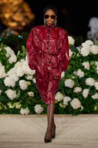 Model walks the runway in a glossy burgundy belted dress with puffed sleeves, oversized orange sunglasses, and bold earrings, against a white floral backdrop.