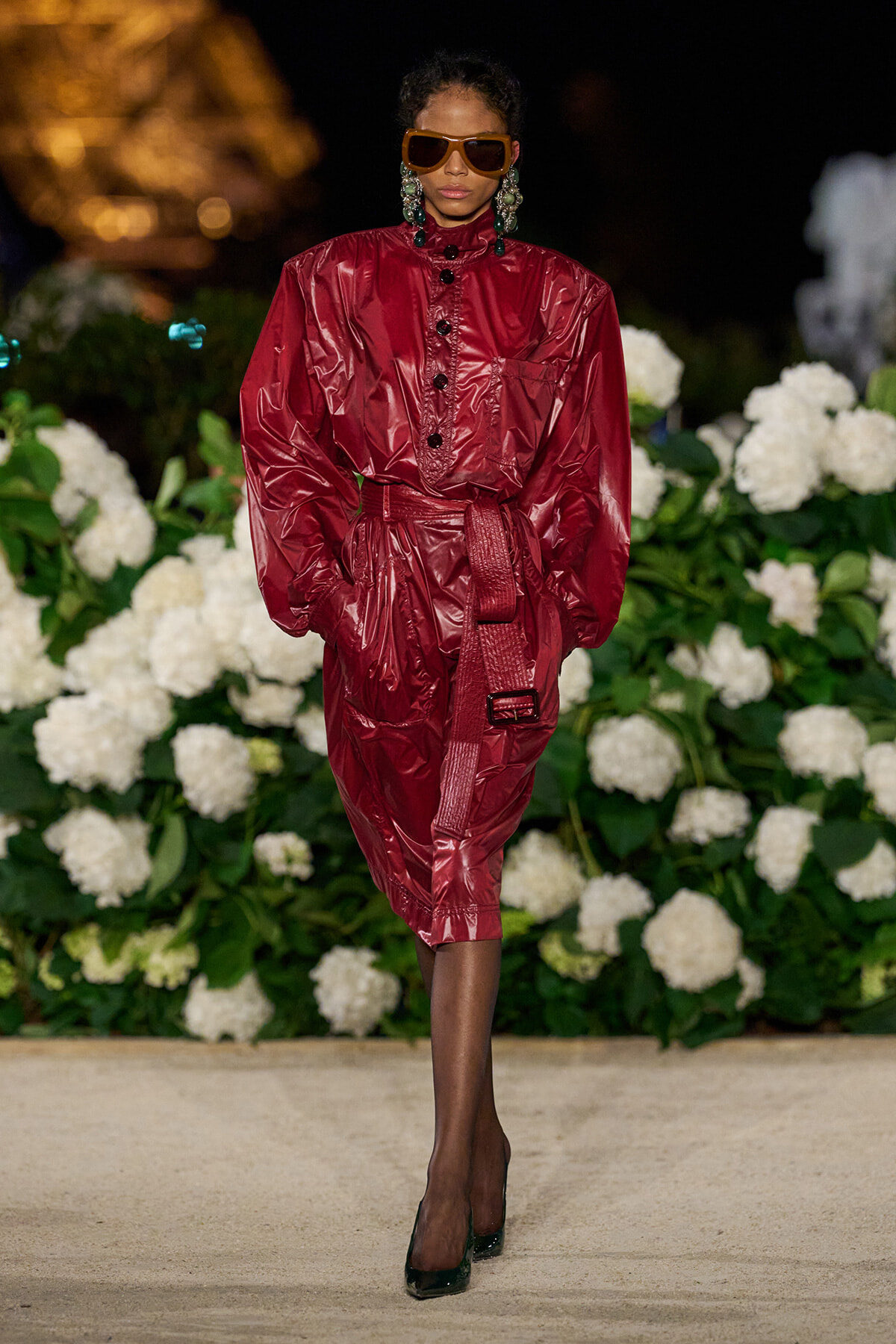Model walks the runway in a glossy burgundy belted dress with puffed sleeves, oversized orange sunglasses, and bold earrings, against a white floral backdrop.