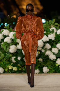 Model walking a runway in a metallic copper-orange coat-dress with a wide belt, oversized shoulders, black sunglasses, and statement earrings, against white hydrangea backdrop.