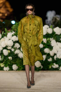 Model in a long olive-green belted trench coat with oversized pockets, walking the runway at night with white hydrangea backdrop and large sunglasses.