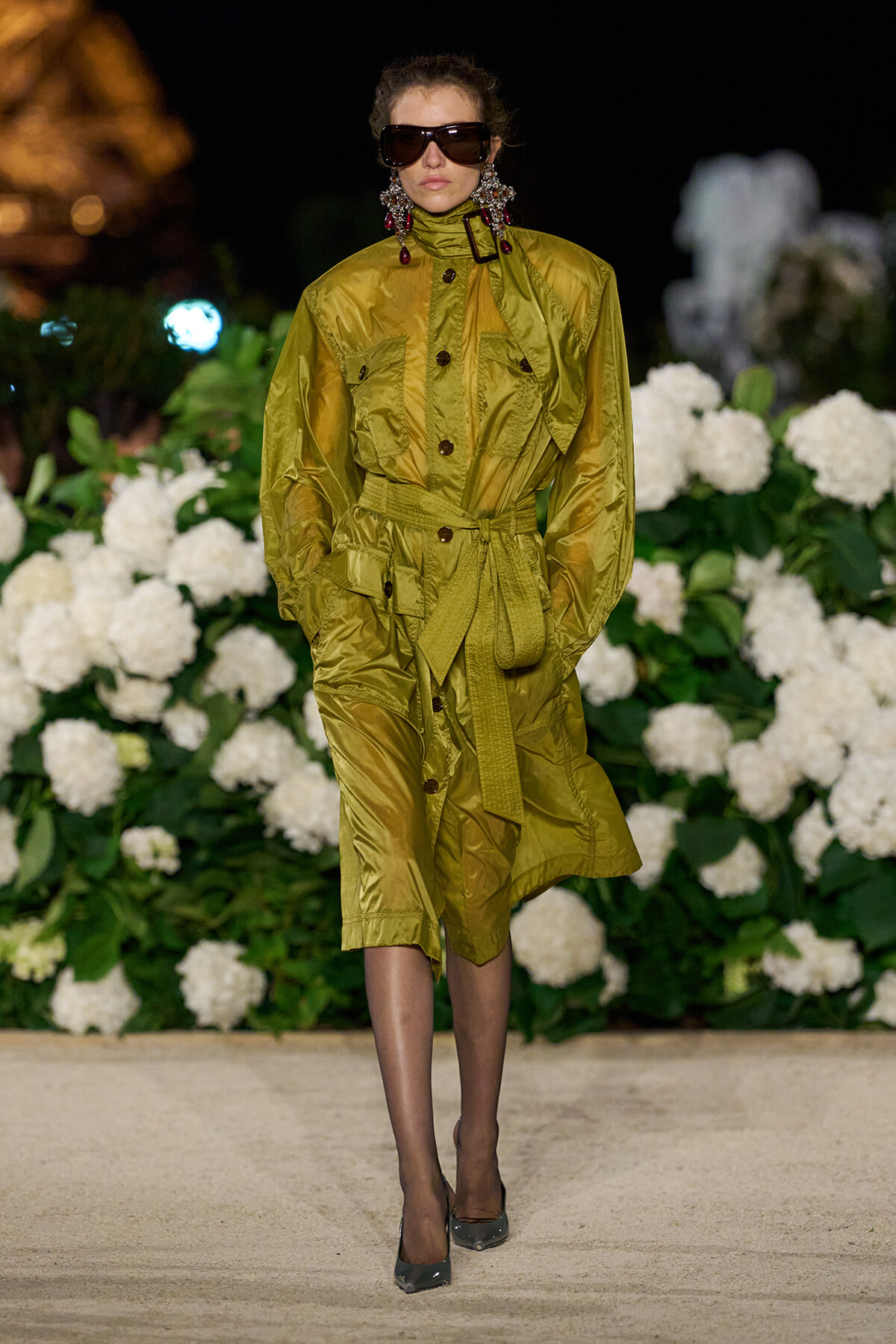 Model in a long olive-green belted trench coat with oversized pockets, walking the runway at night with white hydrangea backdrop and large sunglasses.