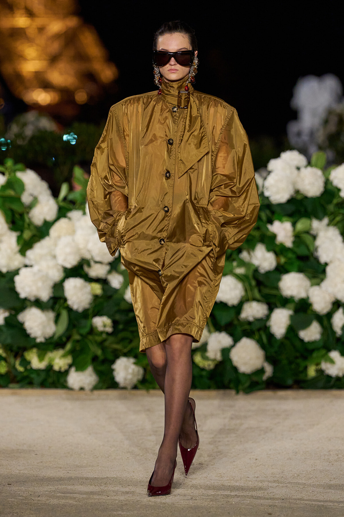 Runway model wearing a shiny oversized gold coat with wide sleeves and red high heels, accessorized with oversized sunglasses and statement earrings, against a backdrop of white hydrangeas at night.