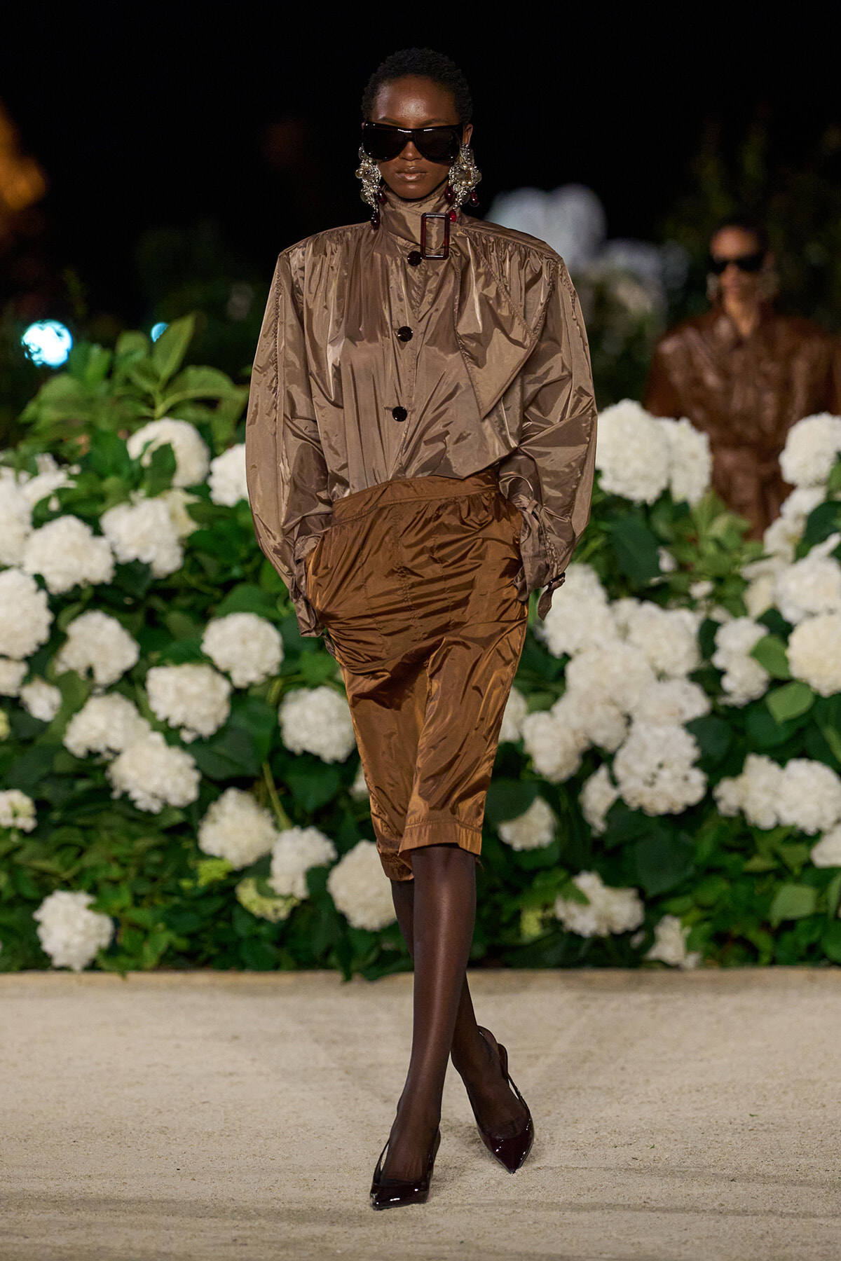 Model walking the runway in a metallic taupe blouse with brown satin culottes, oversized sunglasses, and statement earrings against white florals backdrop.