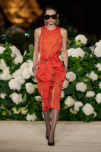 Runway model in an orange sleeveless midi dress with a tied waist, oversized sunglasses, and bold earrings, walking amid white hydrangea backdrop.
