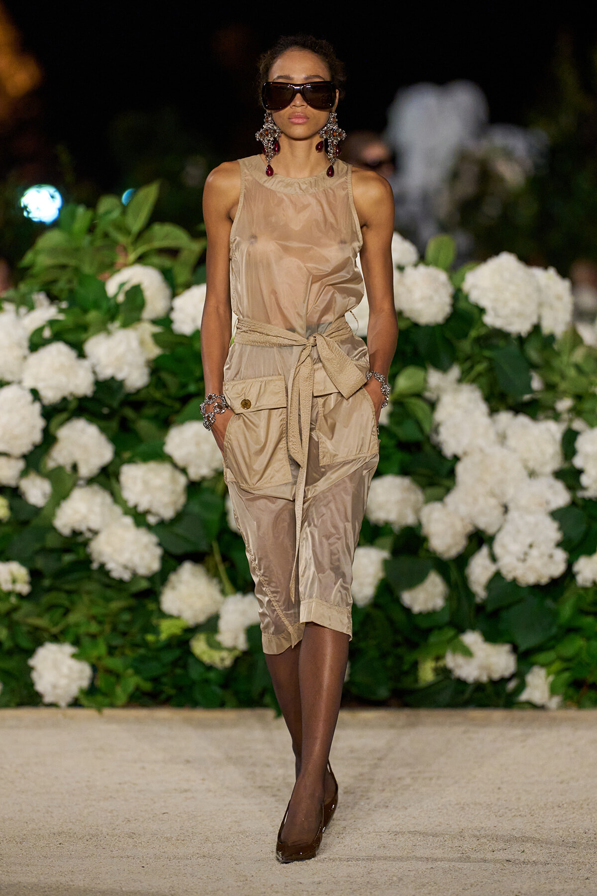 Model walking runway in beige sleeveless jumpsuit with waist tie, statement earrings, and dark sunglasses, against a backdrop of white hydrangeas.