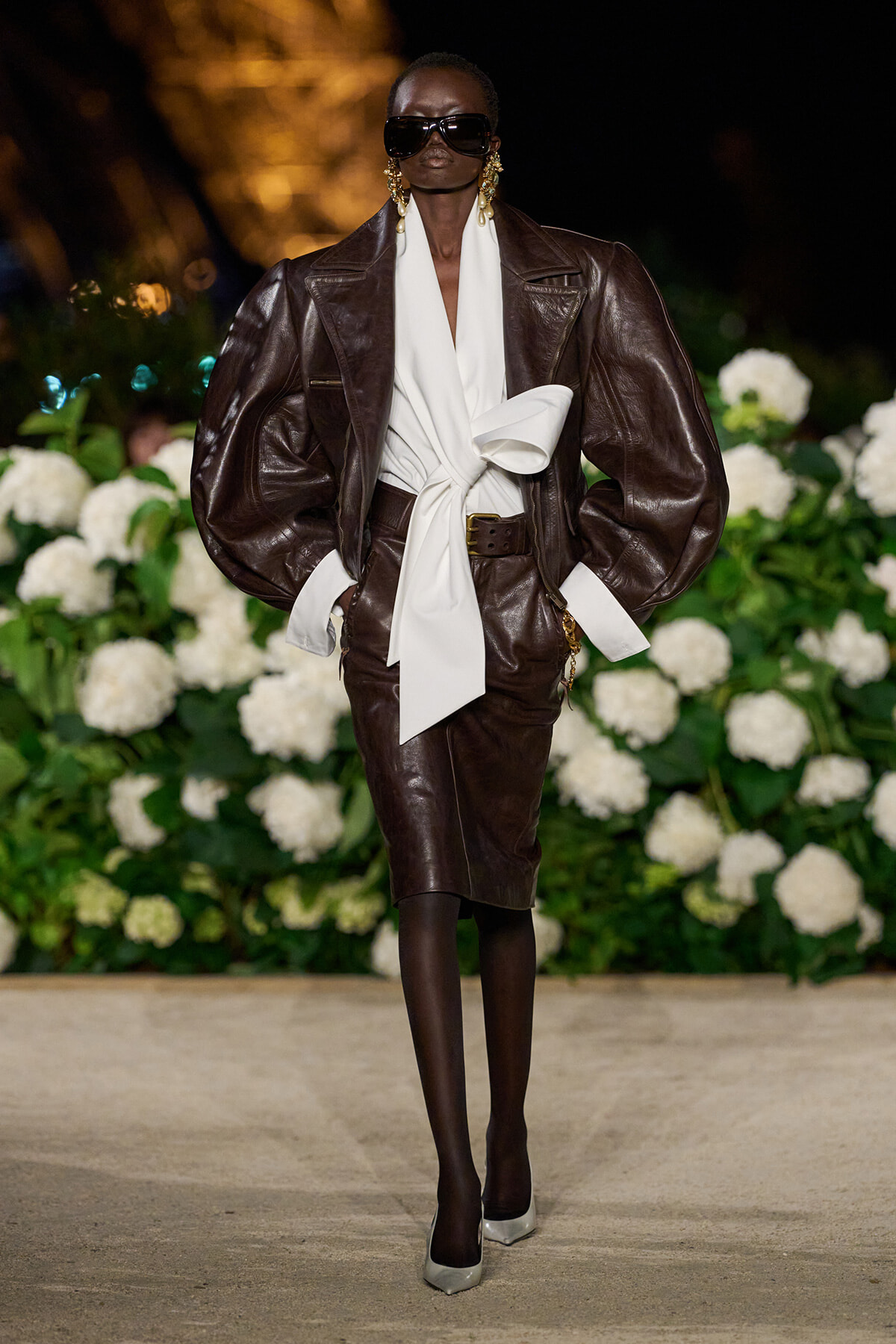 Model on a runway wearing a oversized brown leather suit with a white draped blouse tied in front, large sunglasses, gold earrings, black tights, and silver heels, walks past white hydrangea backdrop.