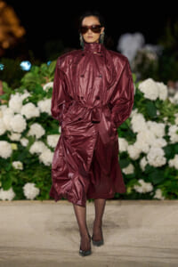 Model walking a runway in a burgundy shiny trench coat belted at the waist, with oversized sunglasses and statement earrings, against a backdrop of white hydrangeas.