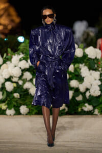 Model on a runway wearing a dark navy shiny trench coat with a belt, oversized sunglasses and bold earrings.