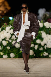 Runway model in a dark brown leather suit with an oversized jacket, white scarf belt, statement earrings, and large sunglasses, walking on a floral backdrop.