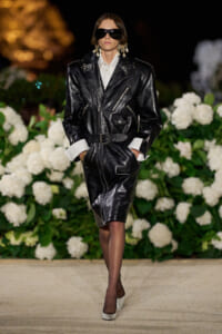 Model walking a runway in a glossy black leather belted jacket with white shirt cuffs, oversized sunglasses, and white heels.