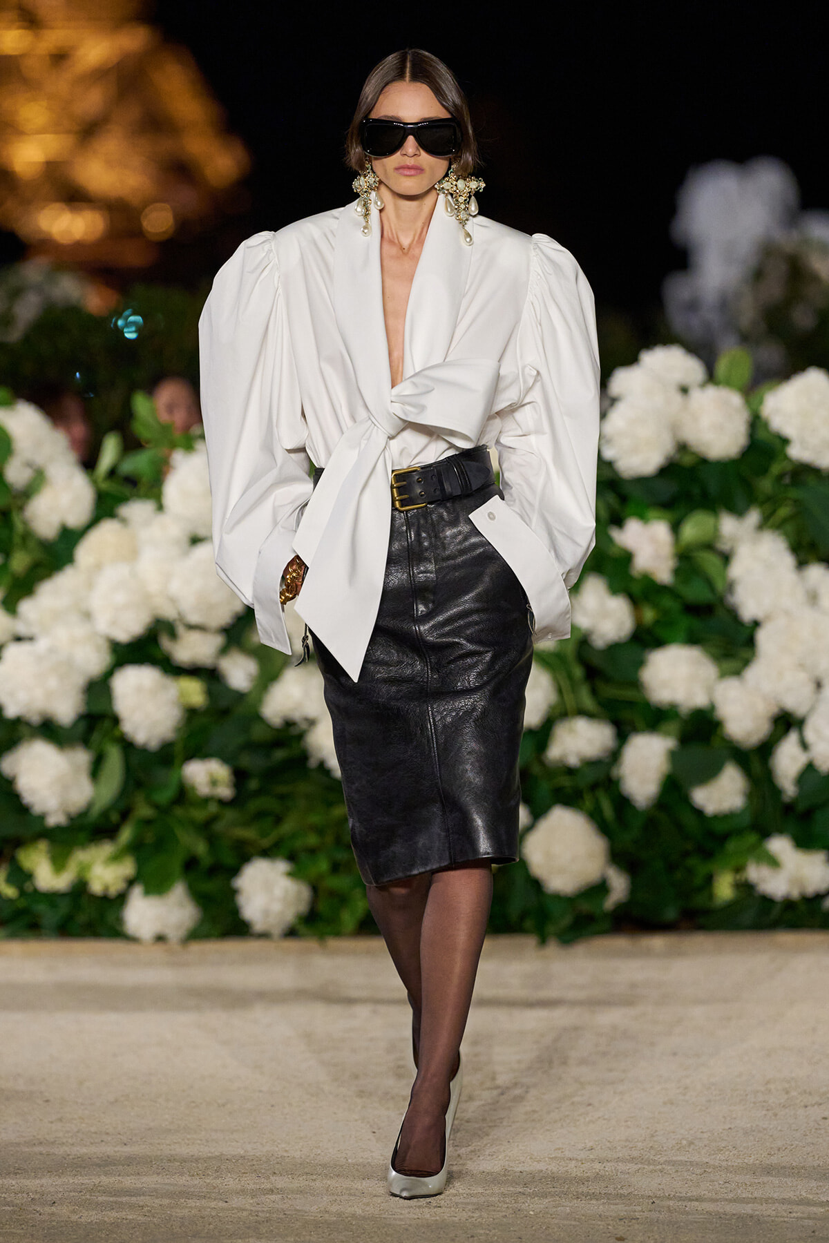 Model on a runway wearing a white oversized blouse tied at the waist with a black leather pencil skirt, a wide belt, large sunglasses and chandelier earrings, white heels, and a floral backdrop.