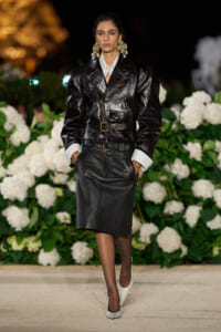 Model walking a black leather belted outfit with a white shirt, oversized jacket, and matching pencil skirt at a night fashion show, surrounded by white hydrangeas.
